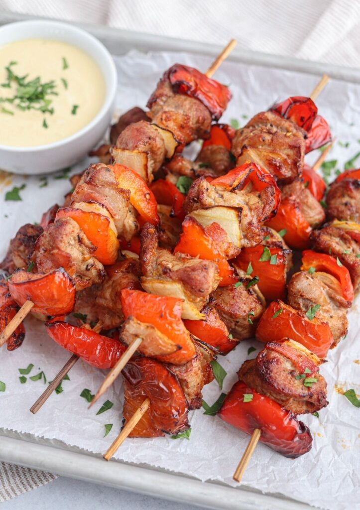 pork kebabs made with peppers, onions and bacon stacked on a baking sheet lined with paper with a side of mustard dipping sauce.