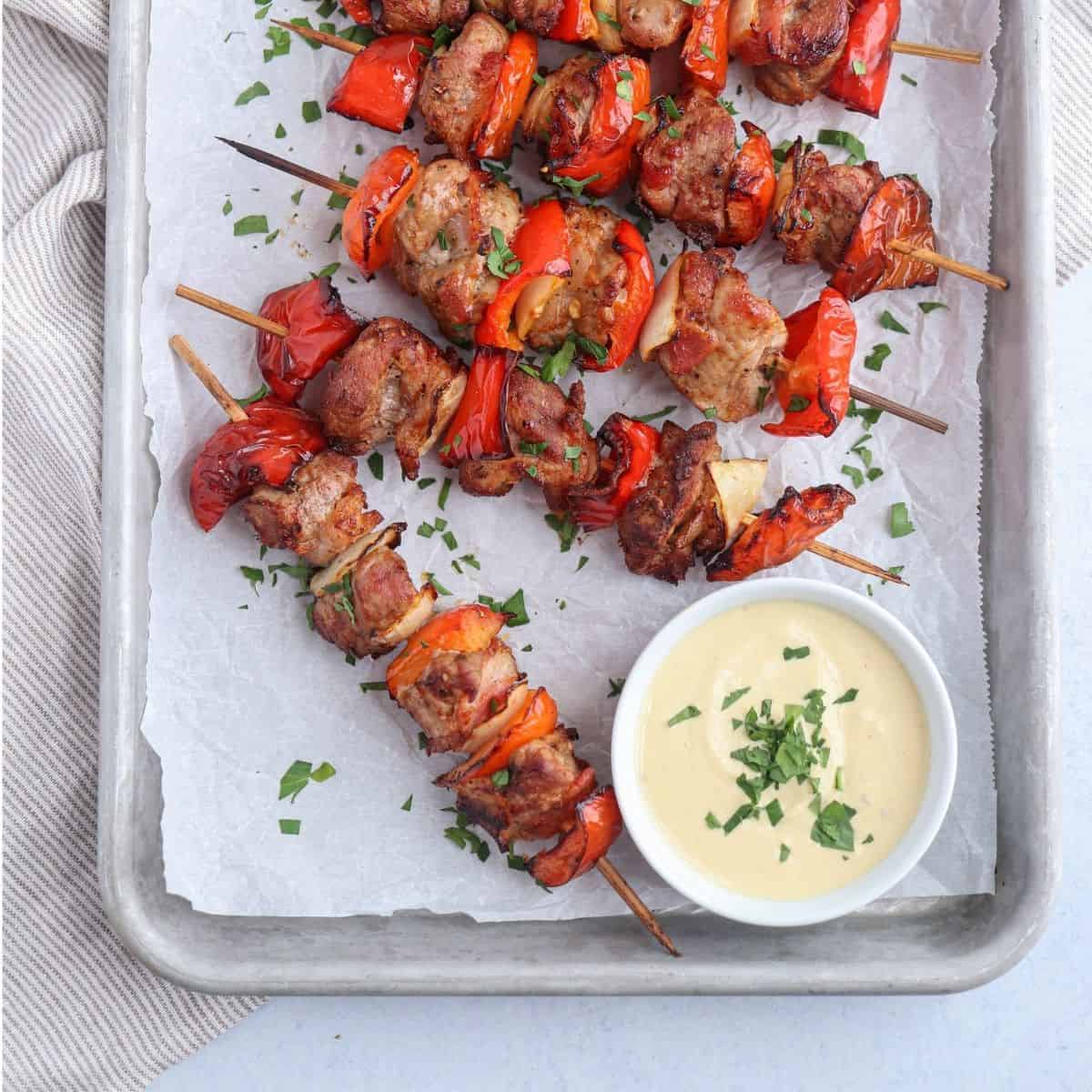 pork kebabs made with peppers, onions and bacon on a baking sheet lined with paper with a side of mustard dipping sauce.