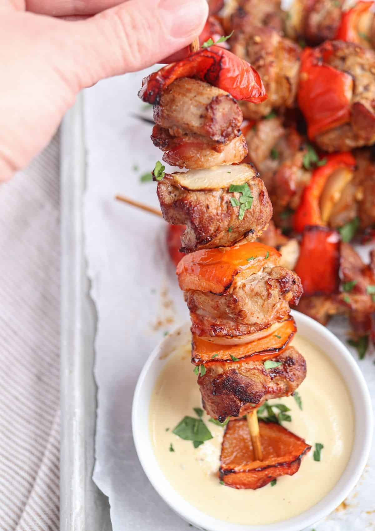 pork shish kebab with red bell pepper, onion and bacon being dipped in creamy mustard sauce.