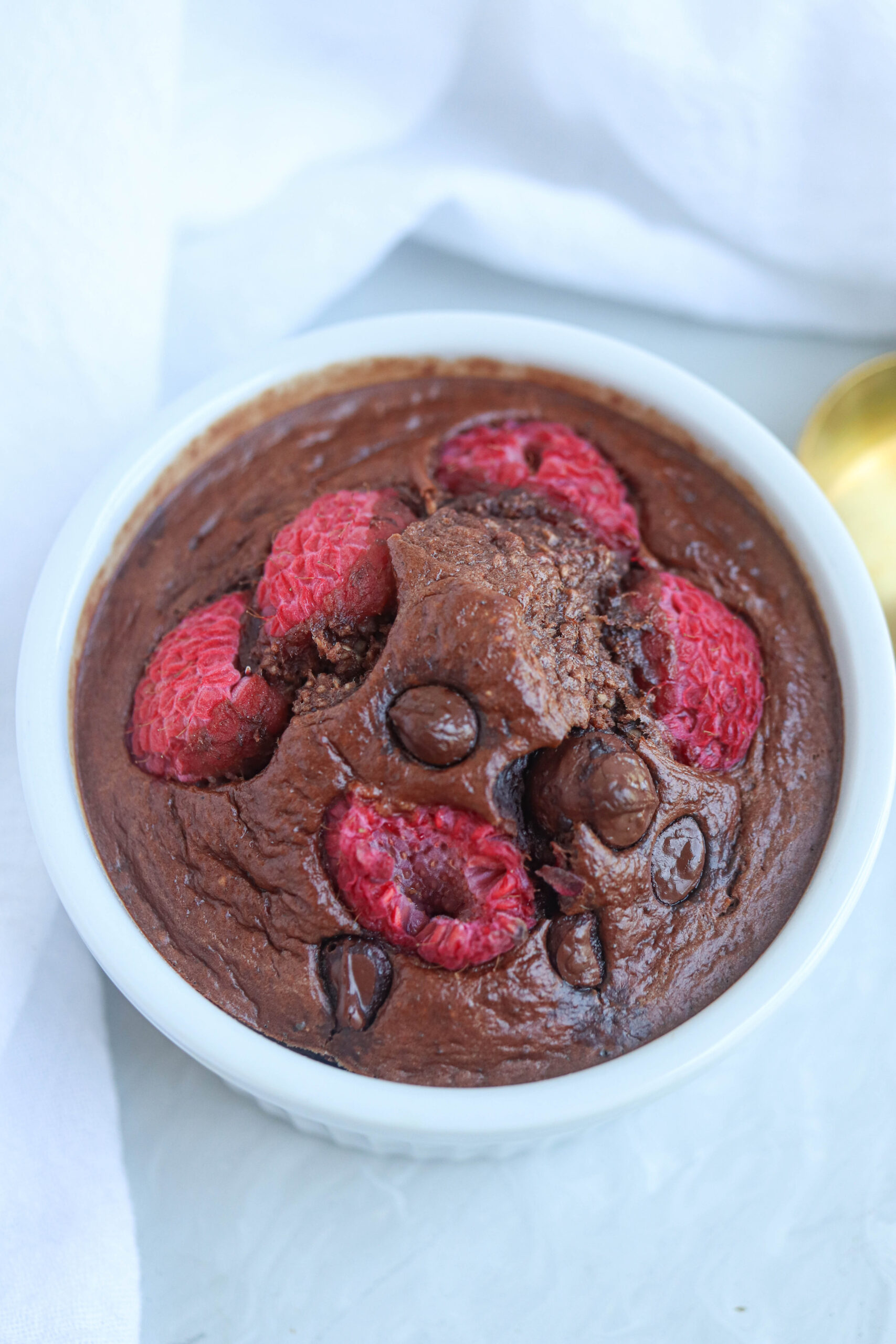 A small white ramekin filled with baked chocolate oatmeal, inspired by healthy banana breakfast recipes, topped with raspberries and chocolate chips, sitting on a light surface with a white cloth nearby.
