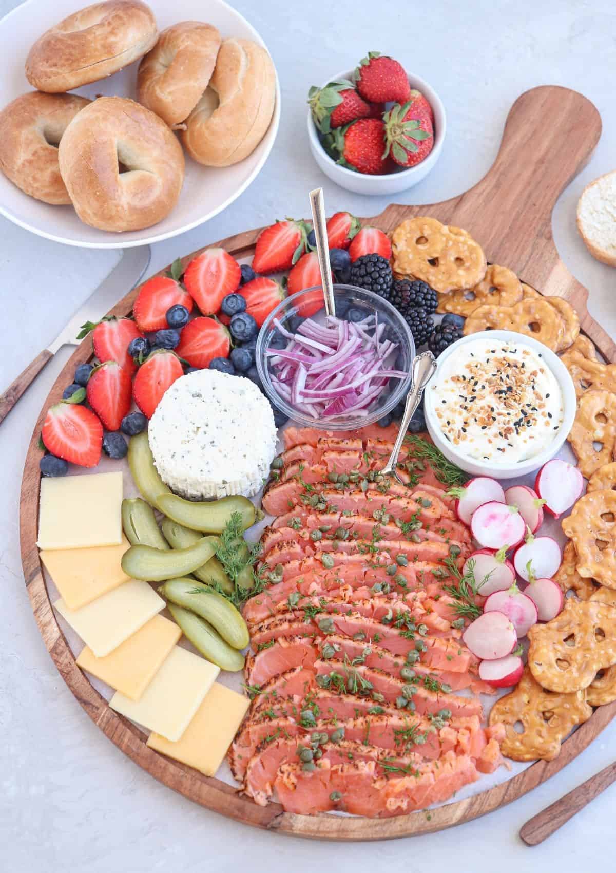 round wooden charcuterie board piled with smoked salmon, fruit, veggies, pickles, pretzel crisps and veggies. 