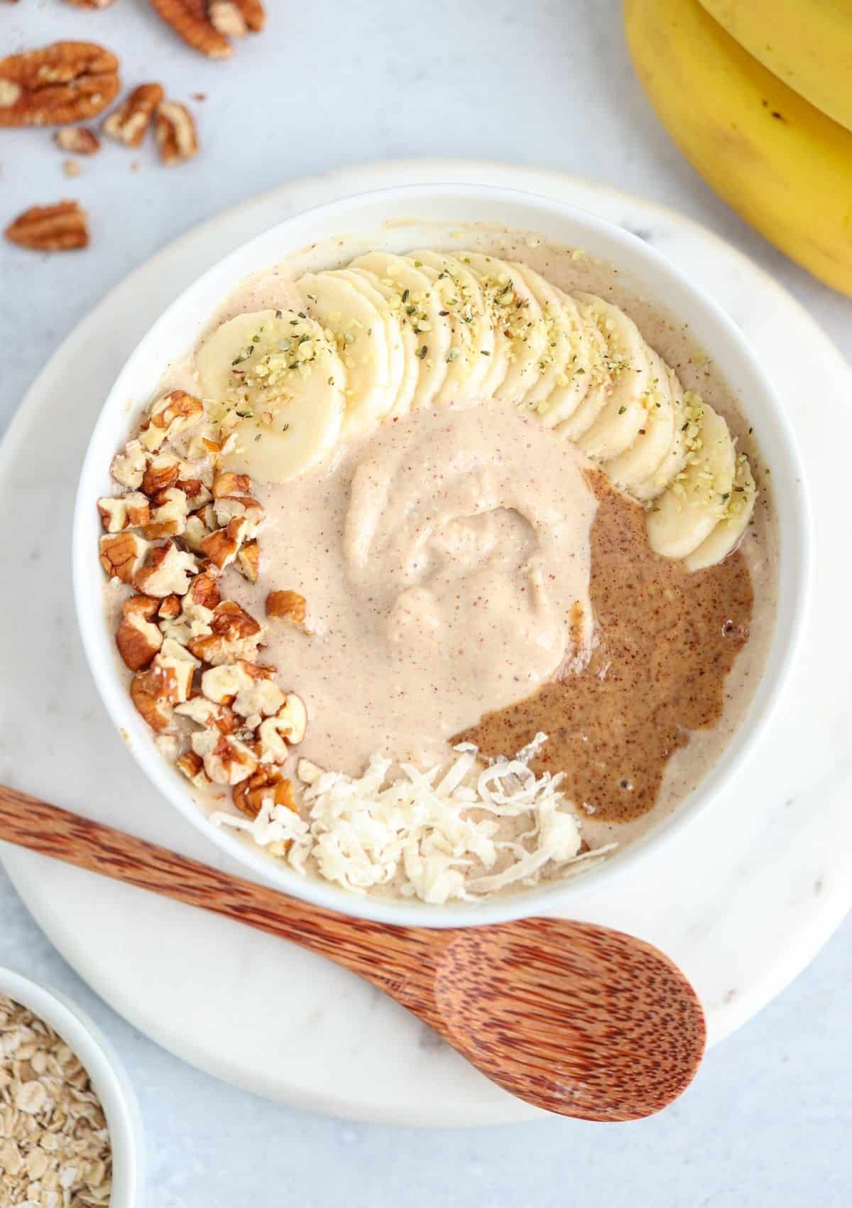 A bowl of oatmeal topped with banana slices, chopped pecans, shredded coconut, nut butter, and hemp seedsโa delicious choice for those seeking healthy banana breakfast recipes. A wooden spoon rests beside the bowl on a white plate.