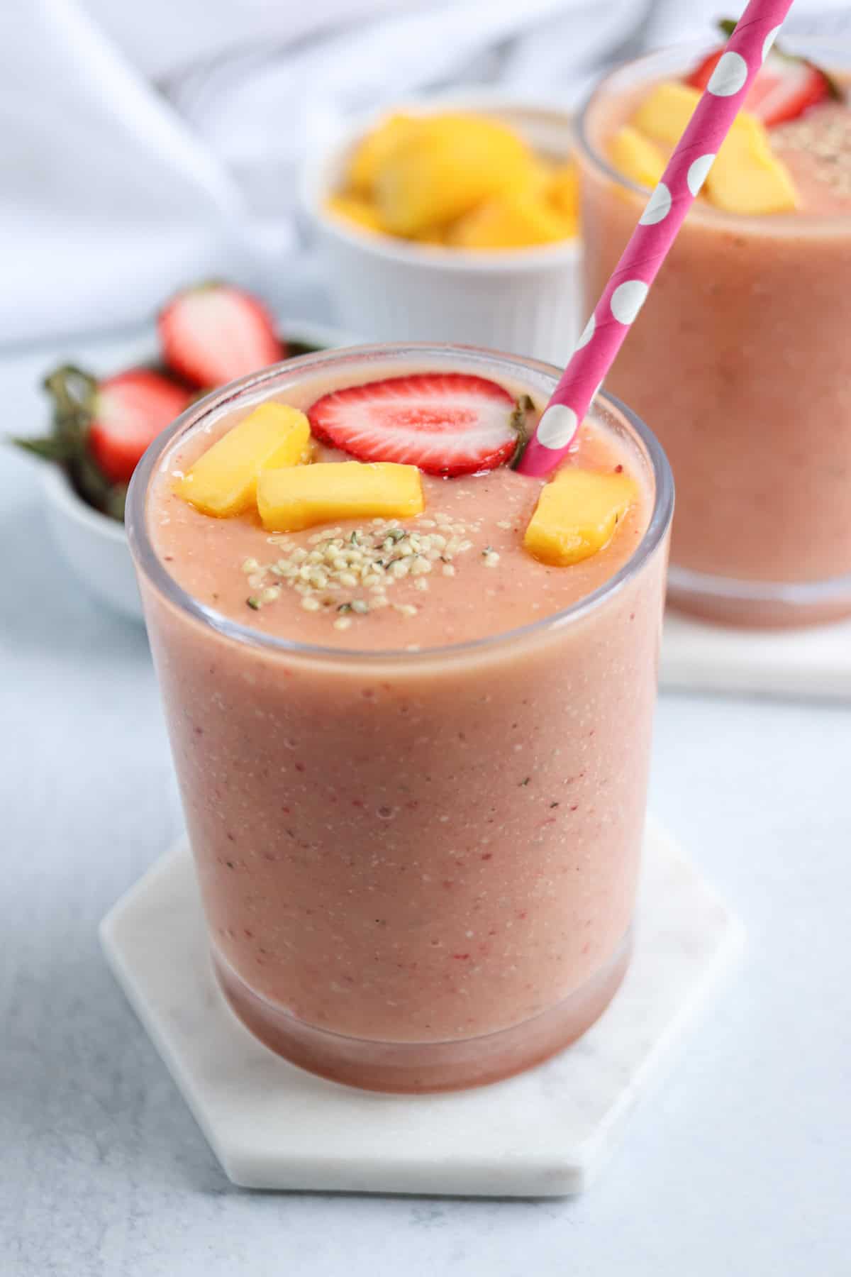 A glass of peach-colored smoothie topped with sliced strawberries, mango chunks, and seedsโperfect for healthy banana breakfast recipes. A pink straw with white polka dots completes the look. Bowls of strawberries and mango sit in the background.