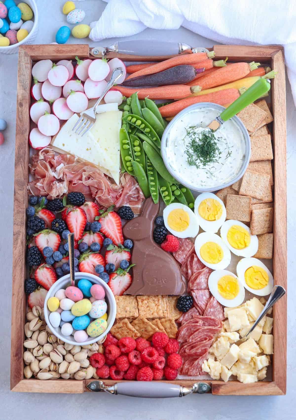 rectangular Easter grazing platter with cured meats, cheese, hard boiled eggs, yogurt dip, fruit, veggies, nuts and candy.