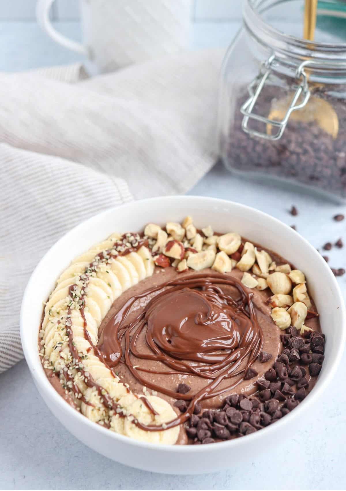 Nutella smoothie bowl in a white bowl topped with sliced banana, melted Nutella, toasted hazelnuts and mini chocolate chips.