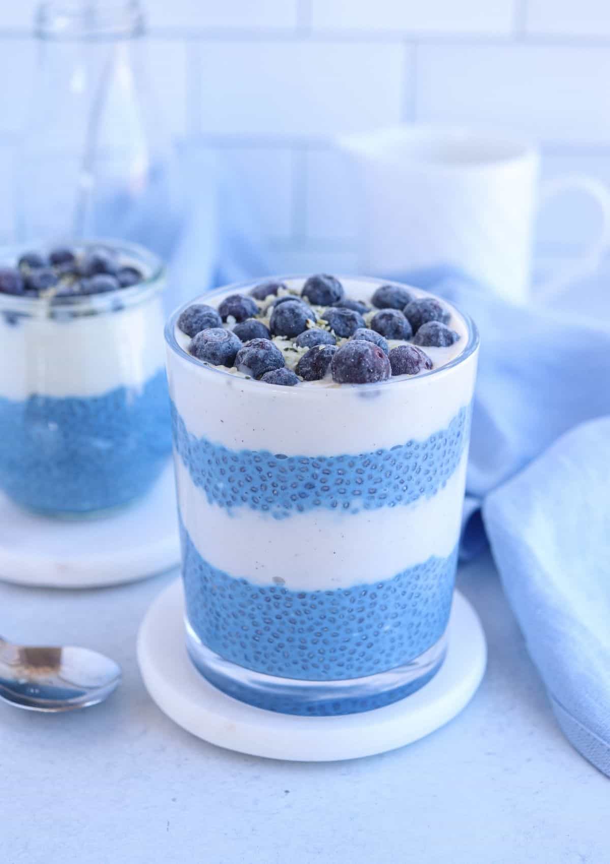 blue chia pudding in a glass layered with yogurt and topped with blueberries and hemp hearts.