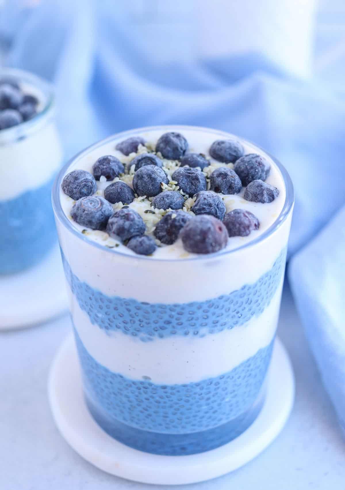 blue chia pudding in a glass layered with yogurt and topped with blueberries and hemp hearts.