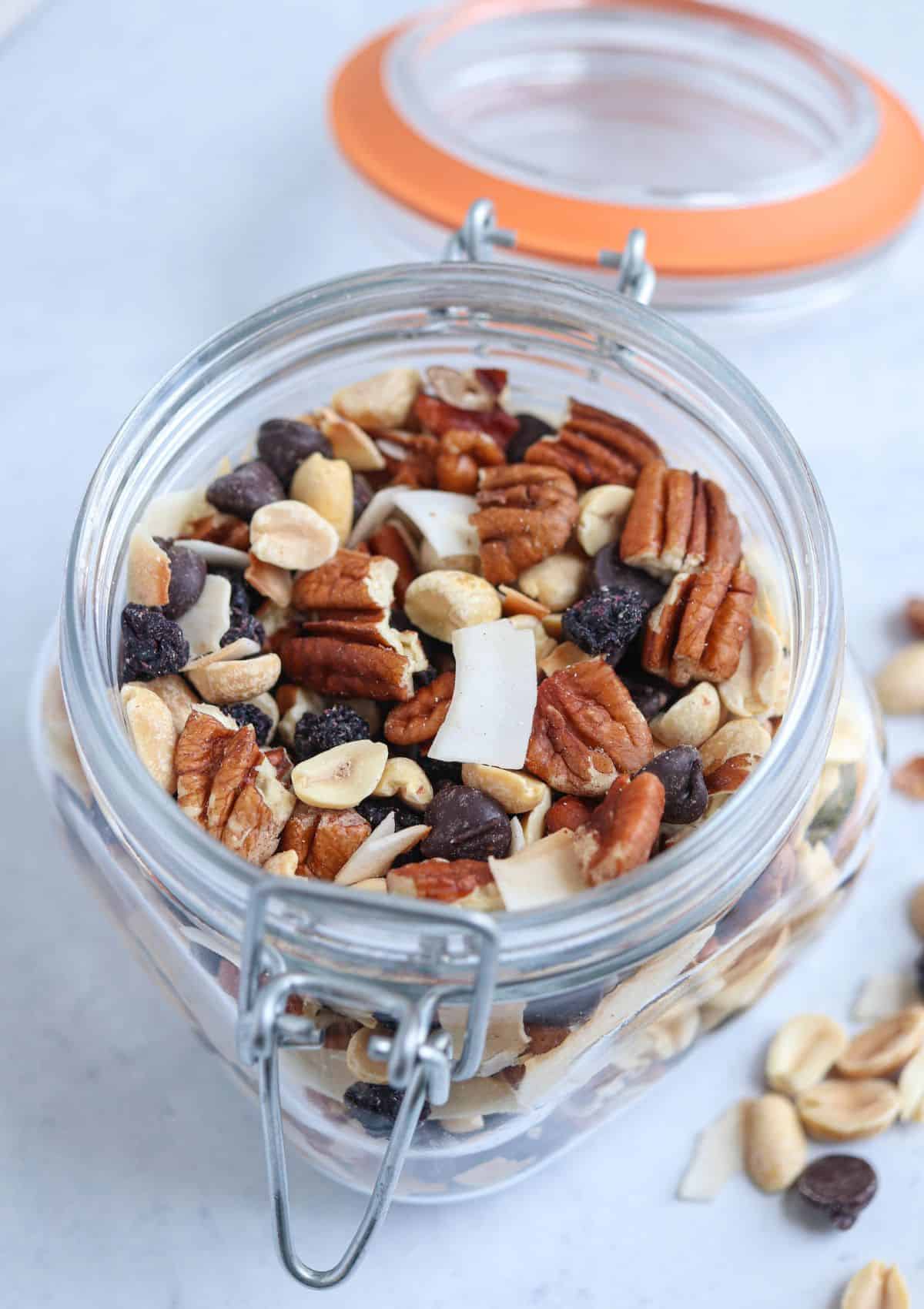 an open jar of low carb trail mix made with nuts, coconut chips, chocolate chips and dried blueberries on a wooden board.