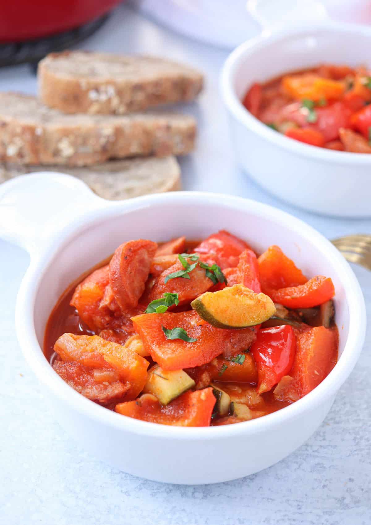 tomato pepper stew with sausage and zucchini in a white bowl with a side of crusty bread.