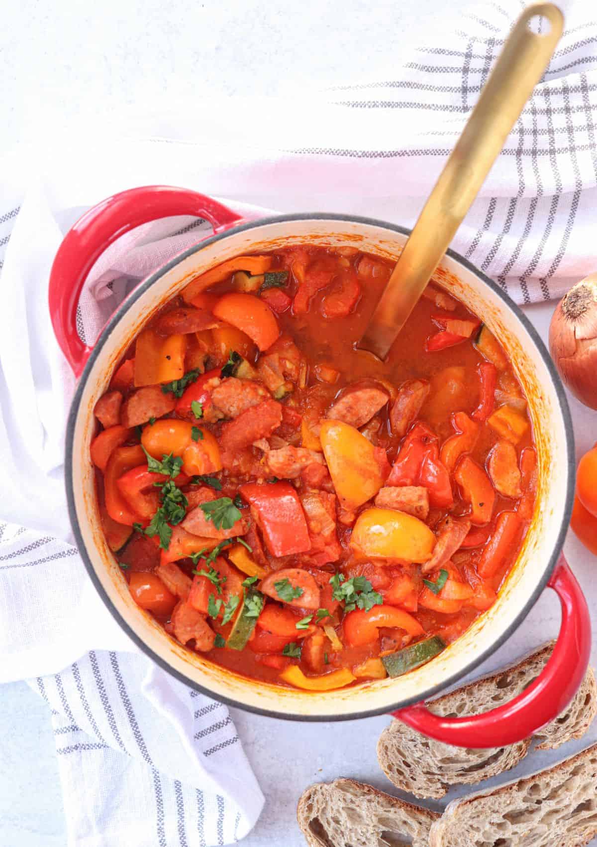 tomato pepper stew with sausage and zucchini in a red Dutch oven with a side of crusty bread.