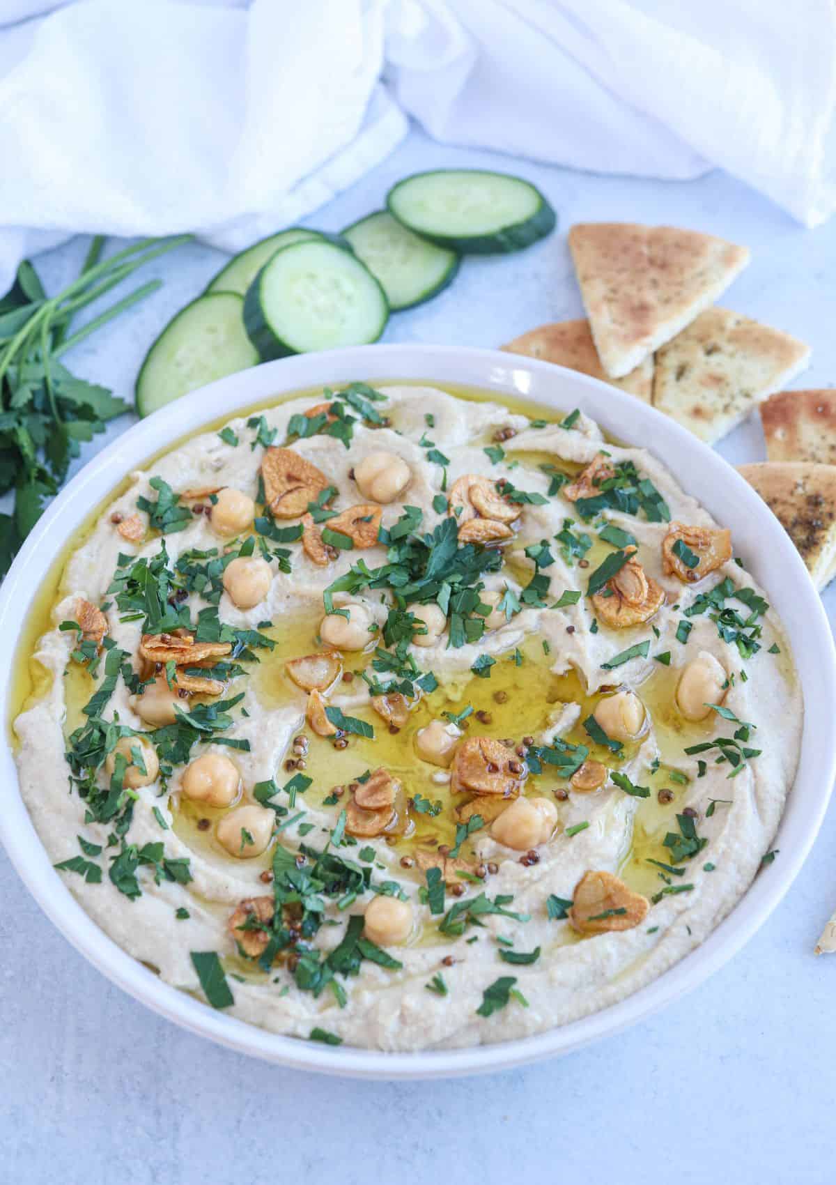 whole chickpea hummus in a white bowl topped with oil, chopped parsley, fried garlic, chickpeas with a side of pita chips, cucumber and parsley.