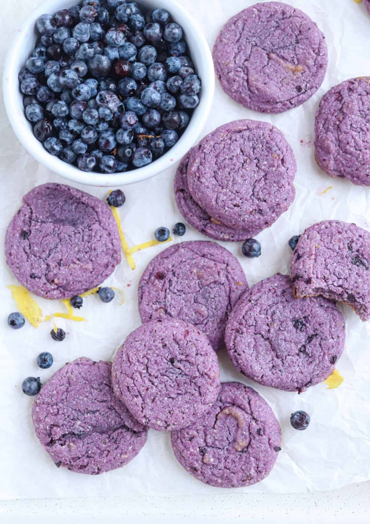 purple blueberry lemon cookies on white parchment paper with blueberries and lemon zest around.