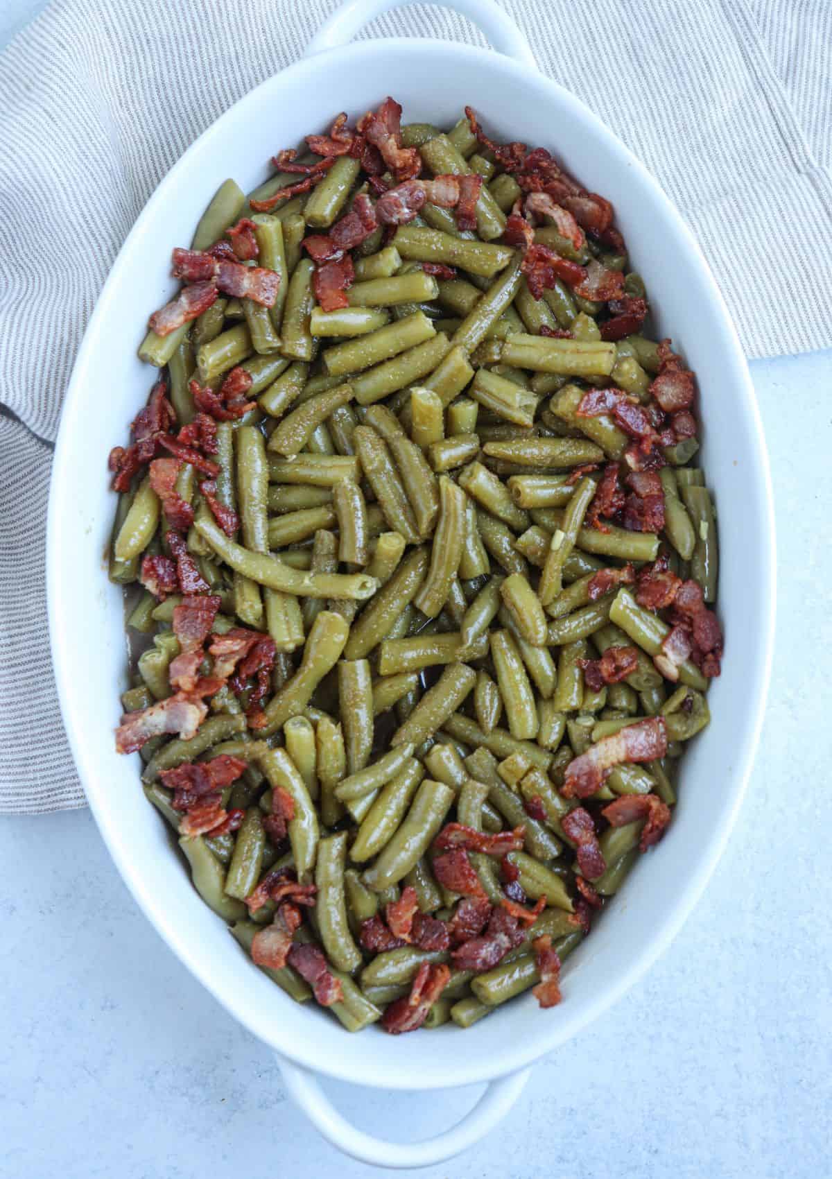 crack green beans in a white oval casserole dish topped with bacon bits on light gray surface. 