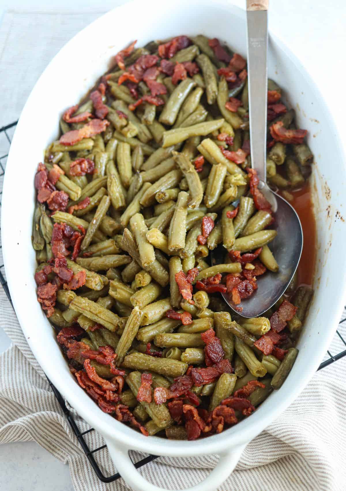 crack green beans in a white oval casserole dish topped with bacon bits  with a large silver spoon in on light gray surface.