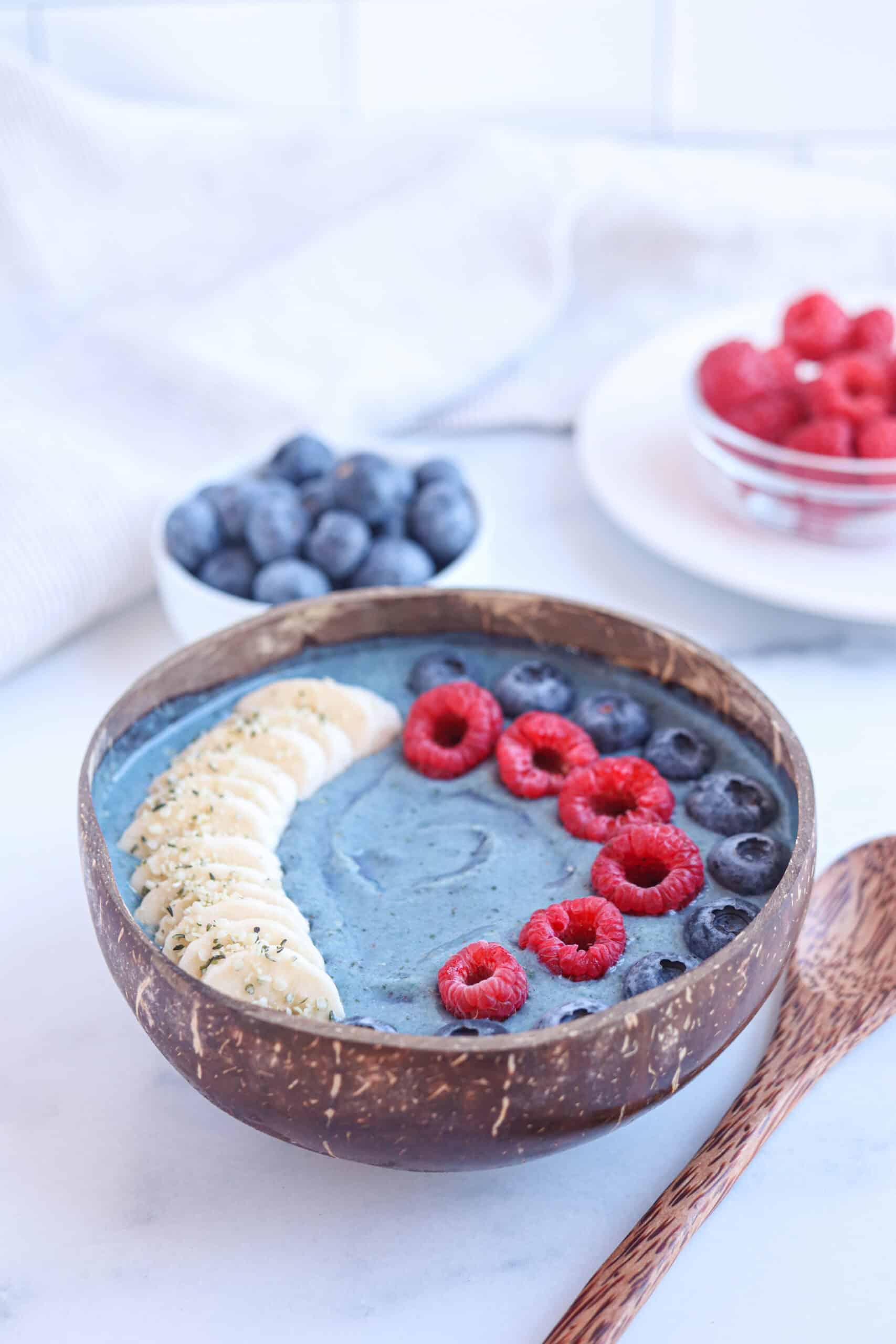 blue spirulina smoothie bowl in a coconut bowl topped with sliced banana, raspberries and blueberries.