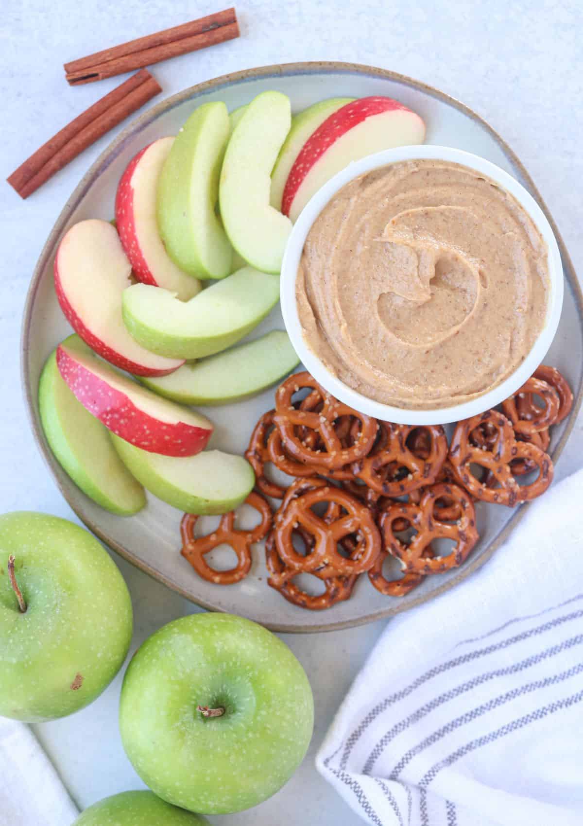 healthy yogurt caramel dip in a white ramekin on a plate surrounded by apple slices and pretzels.