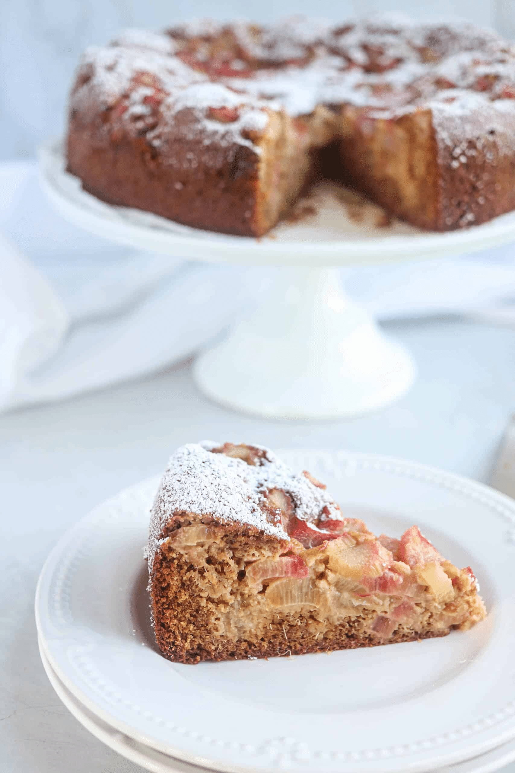 a piece of rhubarb cake on a white plate with the whole cake on a stand in the background. 