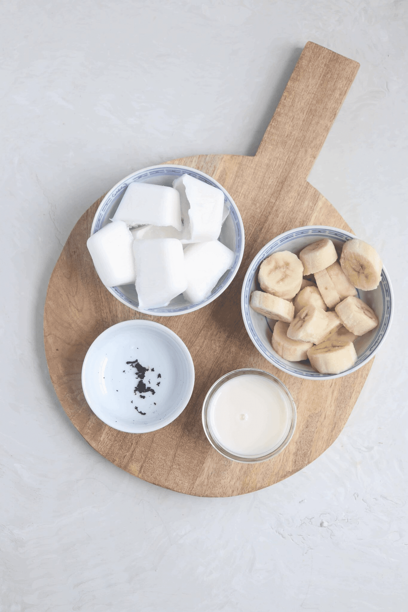 frozen coconut milk, banana, almond milk and vanilla beans in small containers on a round wooden board.