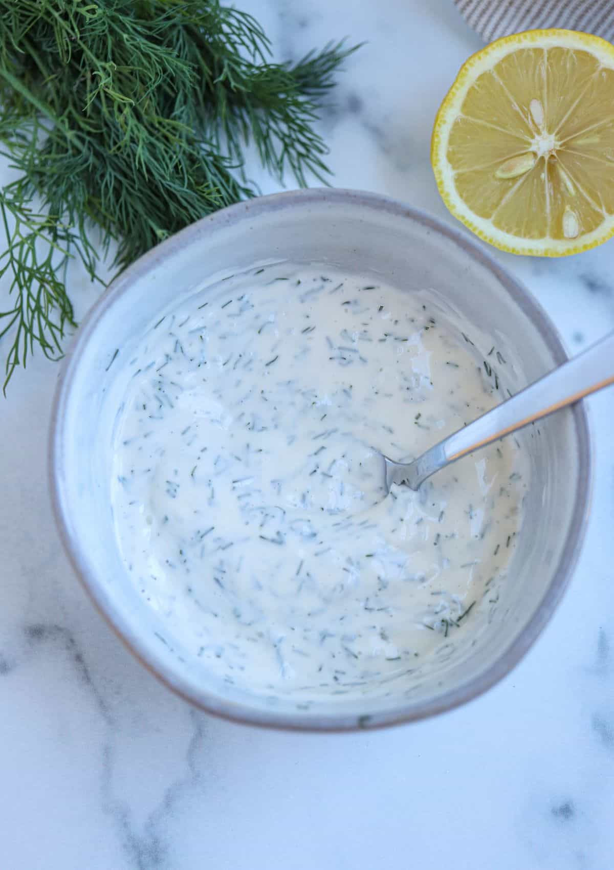 dill aioli mixed with a steel spoon in a medium gray bowl of white marble surface. 