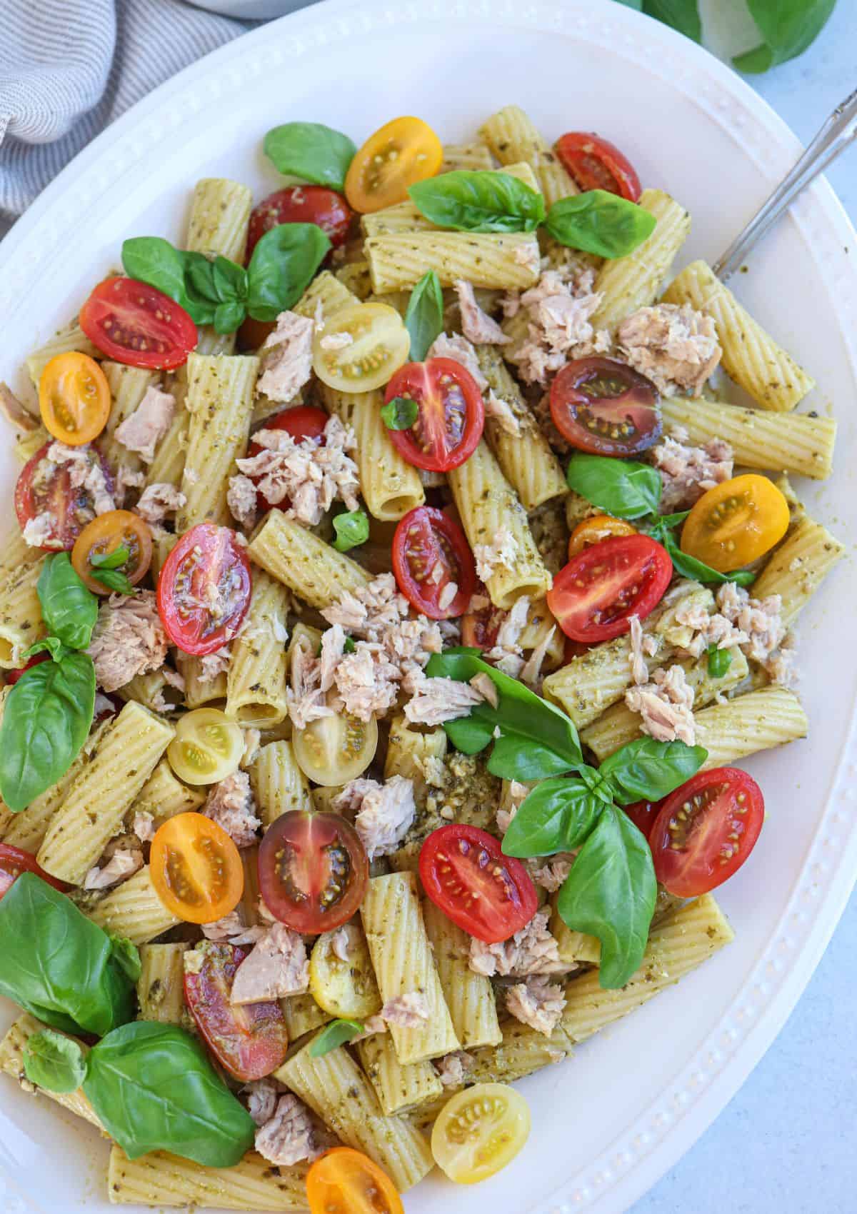 pesto tuna pasta with cherry tomatoes, tuna and fresh basil leaves on an oval white platter.