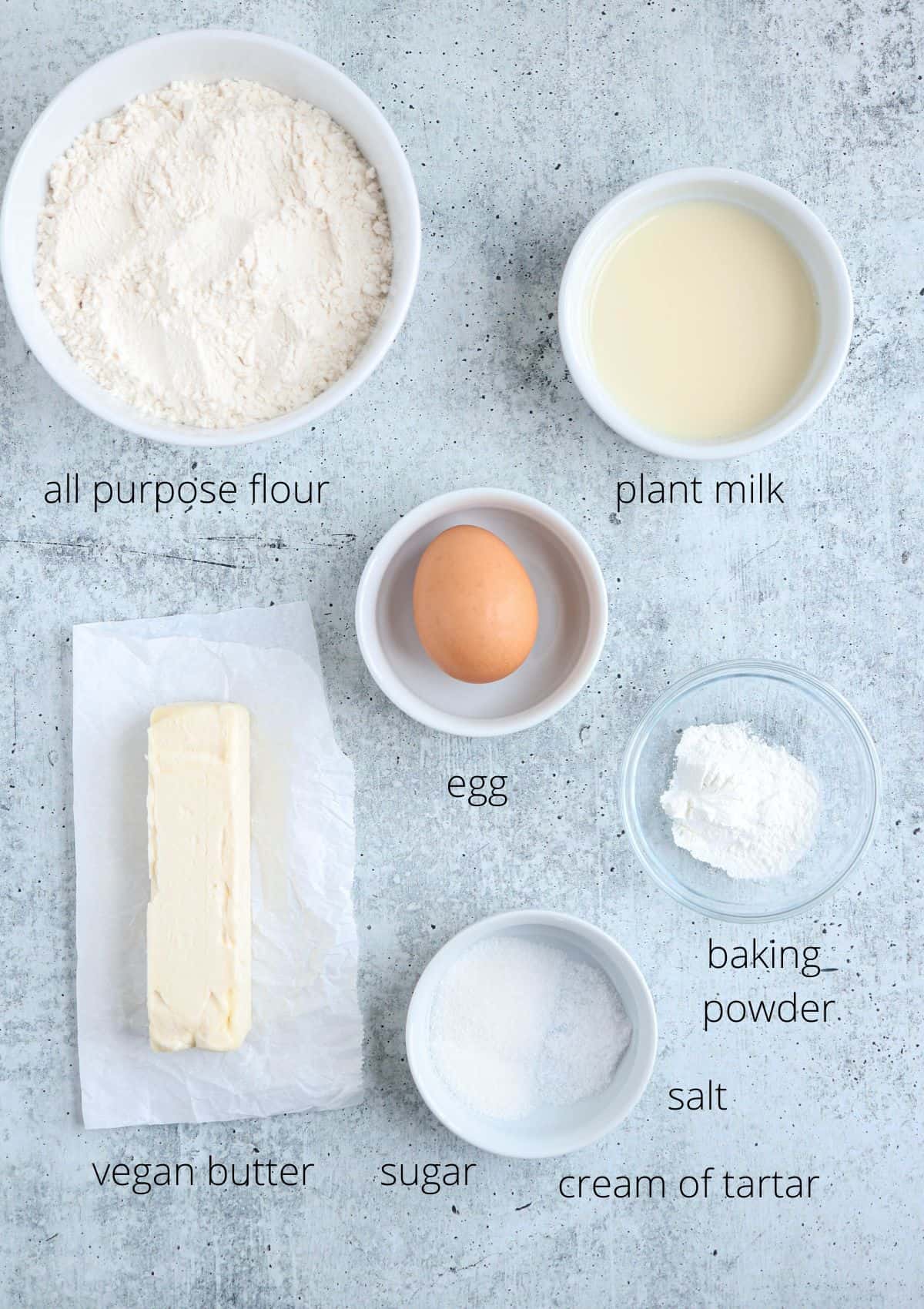 ingredients for making biscuits without milk in round containers on gray surface.