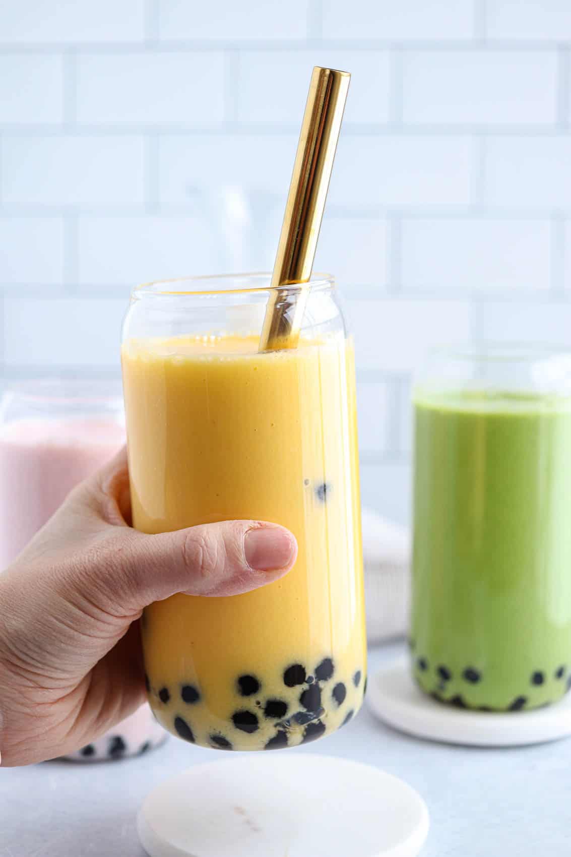 three boba smoothies: yellow, pink and green in beer can shaped glasses, yellow smoothie is held in a hand. 