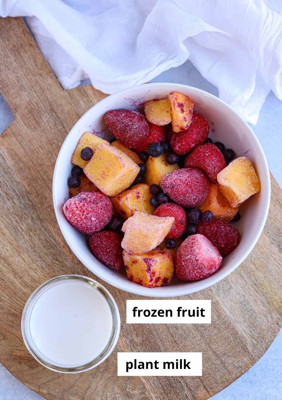 frozen fruit in a white bowl and a glass jar with plant milk on a wooden board.
