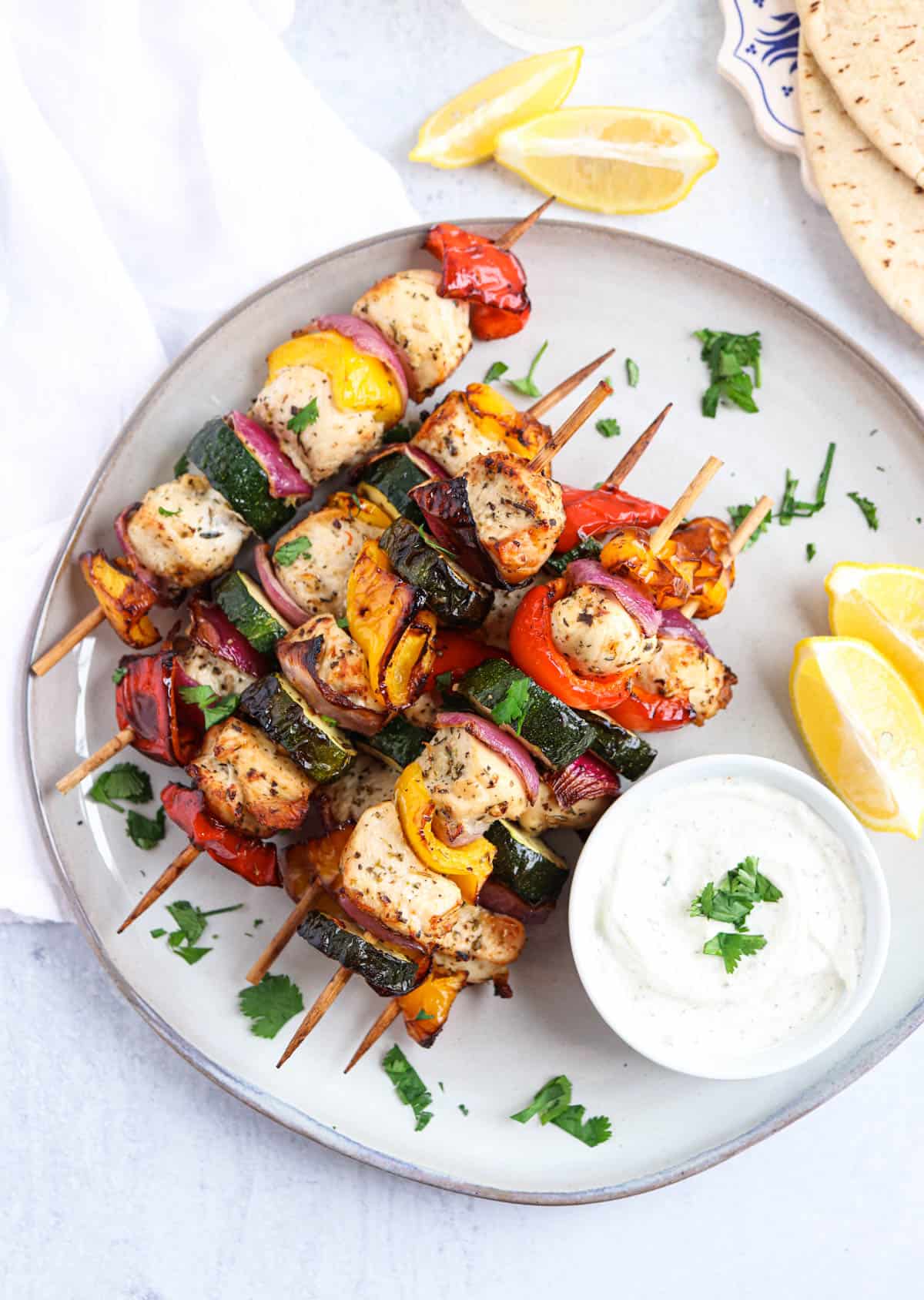 chicken and vegetable kebabs on wooden skewers stacked on a gray plate with a side of ranch dip and lemon wedges.