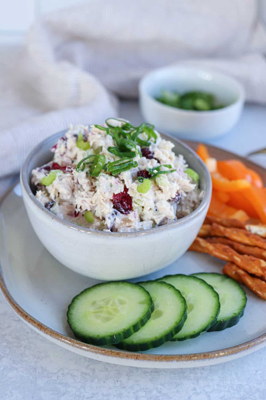 creamy chicken salad in a small gray bowl with chopped scallions on top on a plate with cucumber slices, pretzel sticks and orange bell pepper.