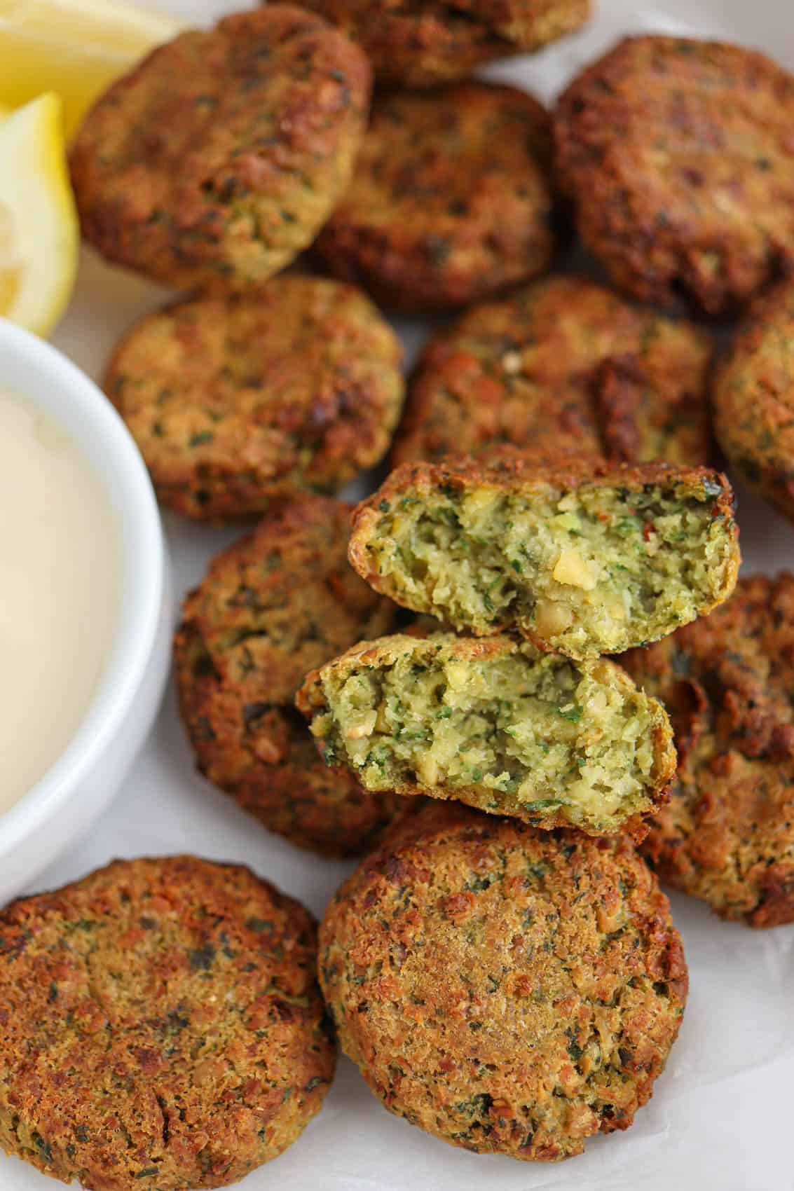 air fried falafel patties on a paper lined place with a side of tahini sauce with one pattie split open to show texture.