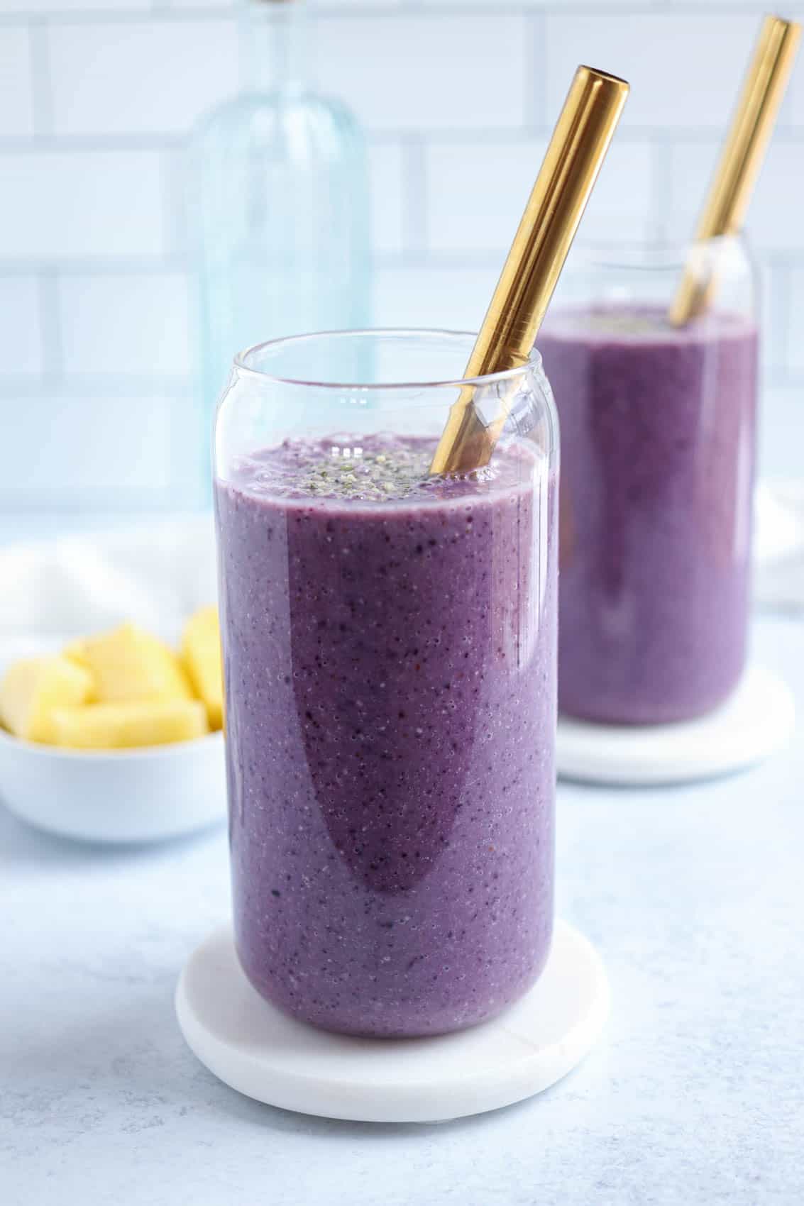 two glasses of blueberry pineapple smoothie with gold straws on light gray surface.