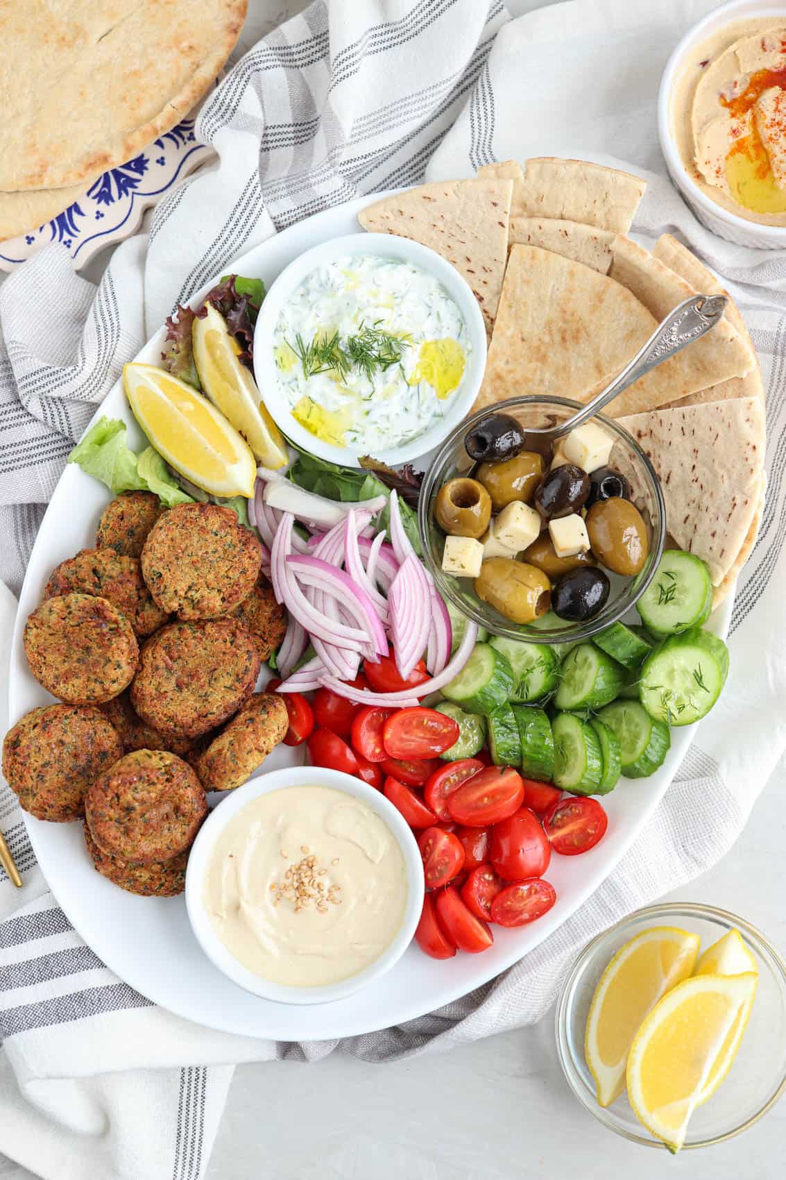 oval falafel platter with falafel patties, pita triangles, fresh veggies, two dips, greens and olives.