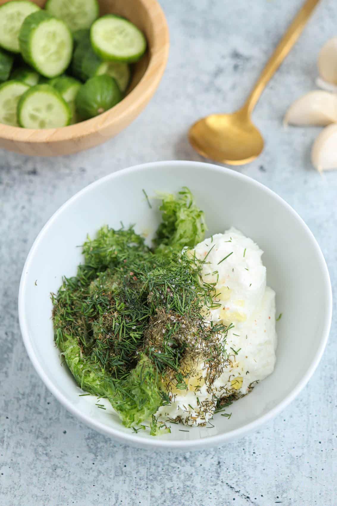 a white bowl with yogurt, grated cucumber, spices and fresh dill before mixing on gray counter.