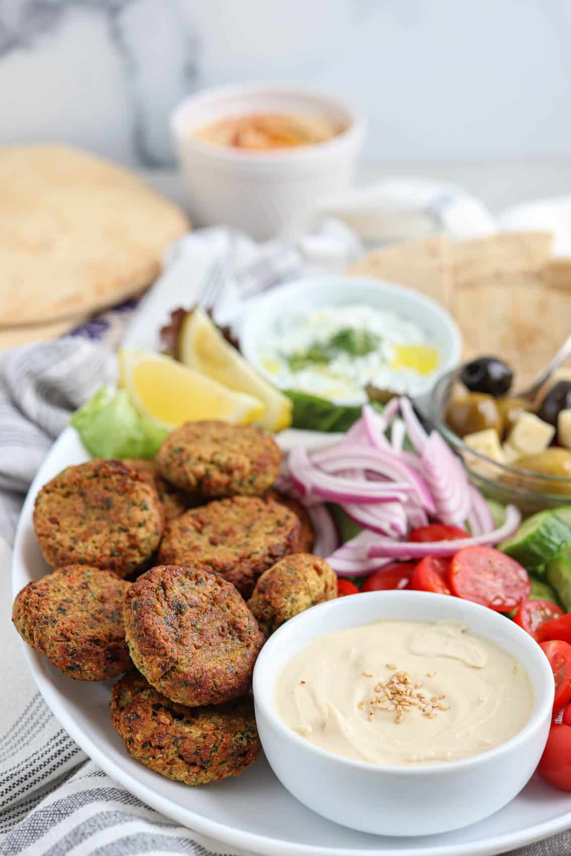 oval falafel platter with falafel patties, pita triangles, fresh veggies, two dips, greens and olives.