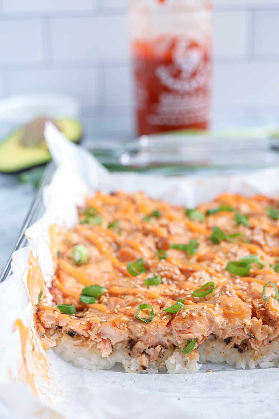 salmon sushi bake topped with sriracha mayo and scallions in a casserole dish.