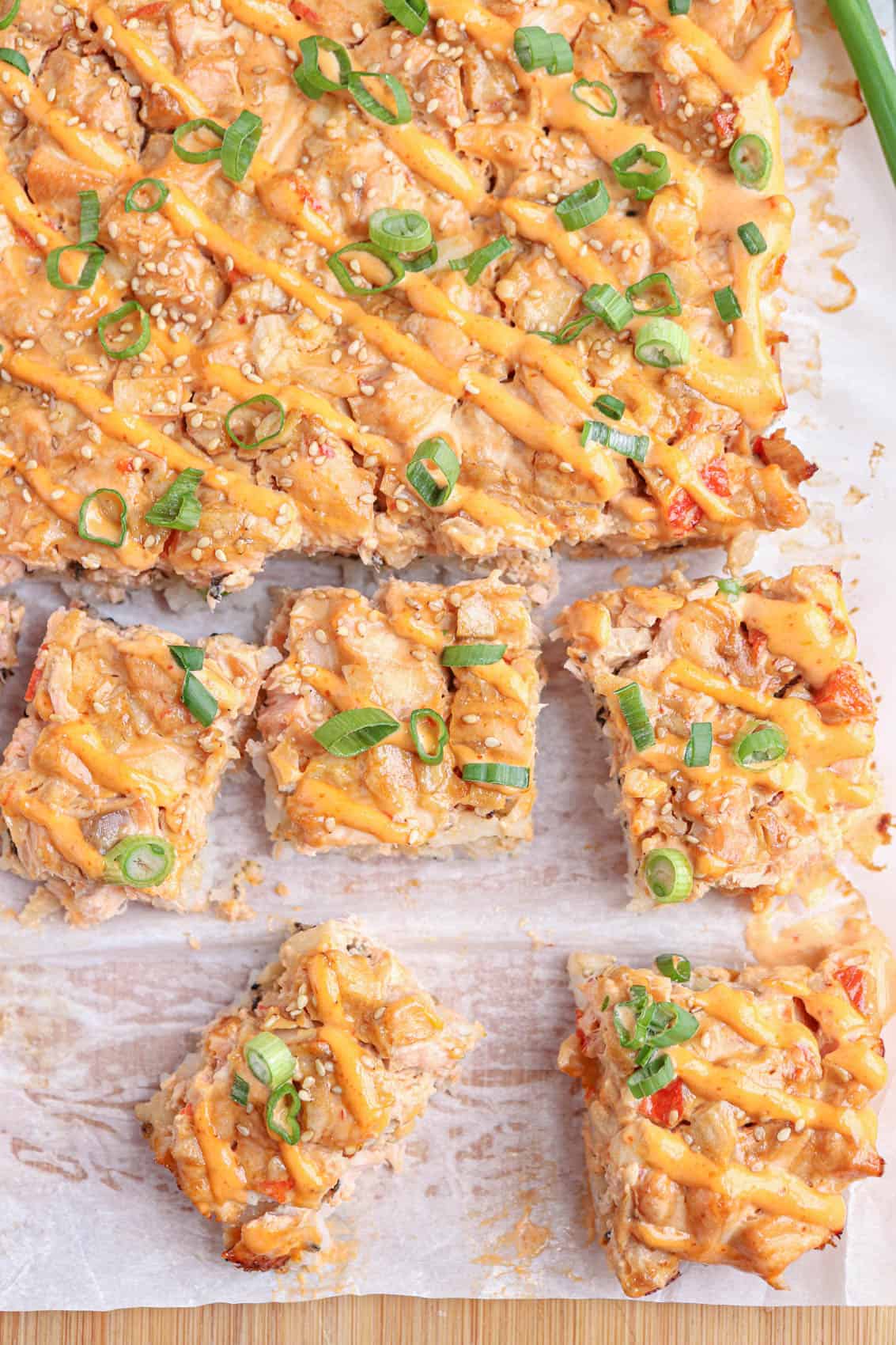 salmon sushi bake topped with sriracha mayo and scallions cut up into small squares.