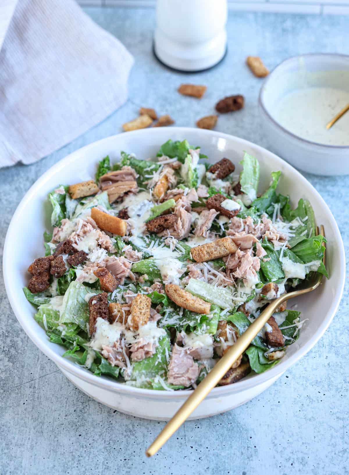 tuna caesar salad with croutons in a white bowl with a golden spoon.