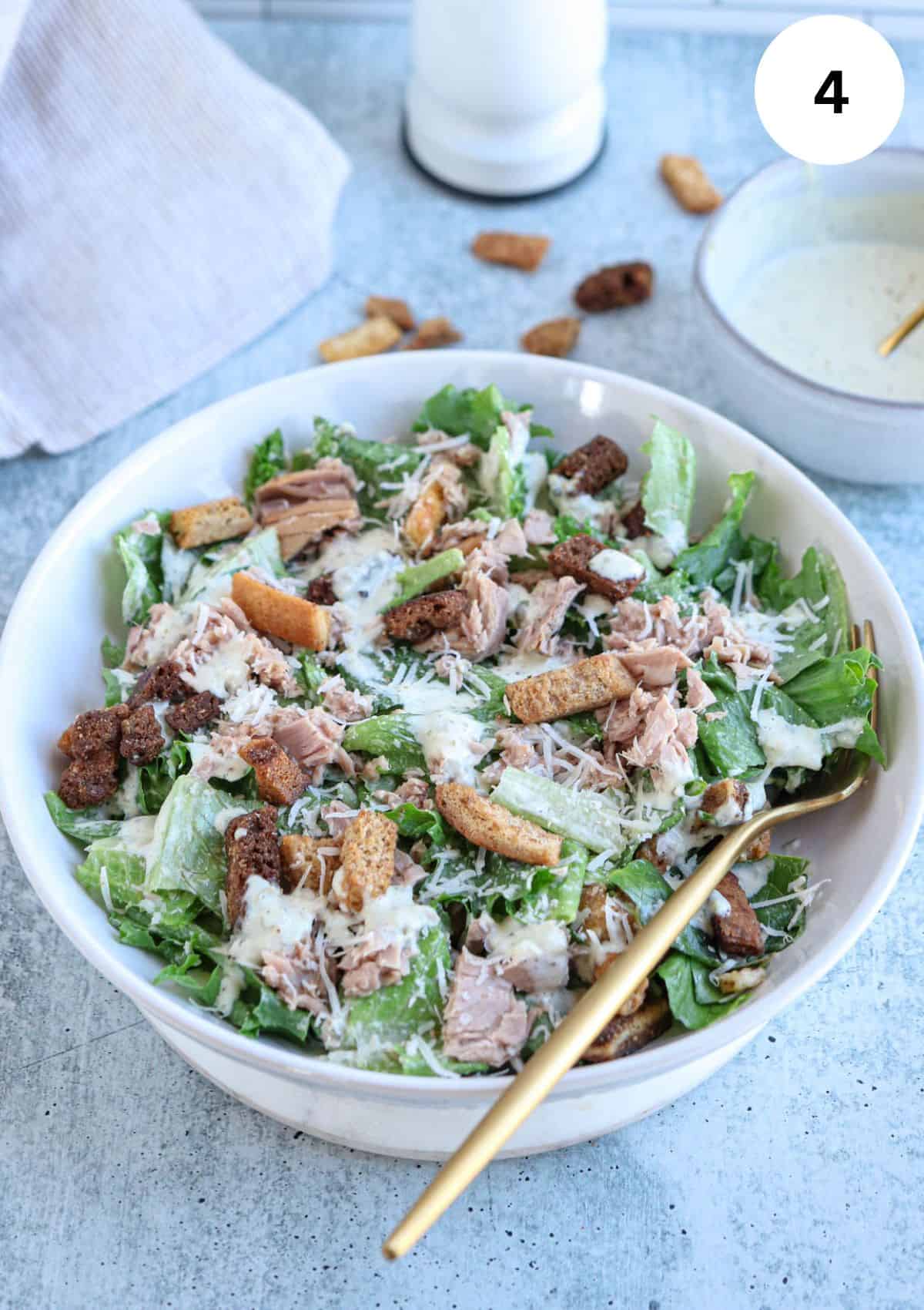 caesar salad wth coutons , tuna, parmesan cheese and dressing in a white bowl.