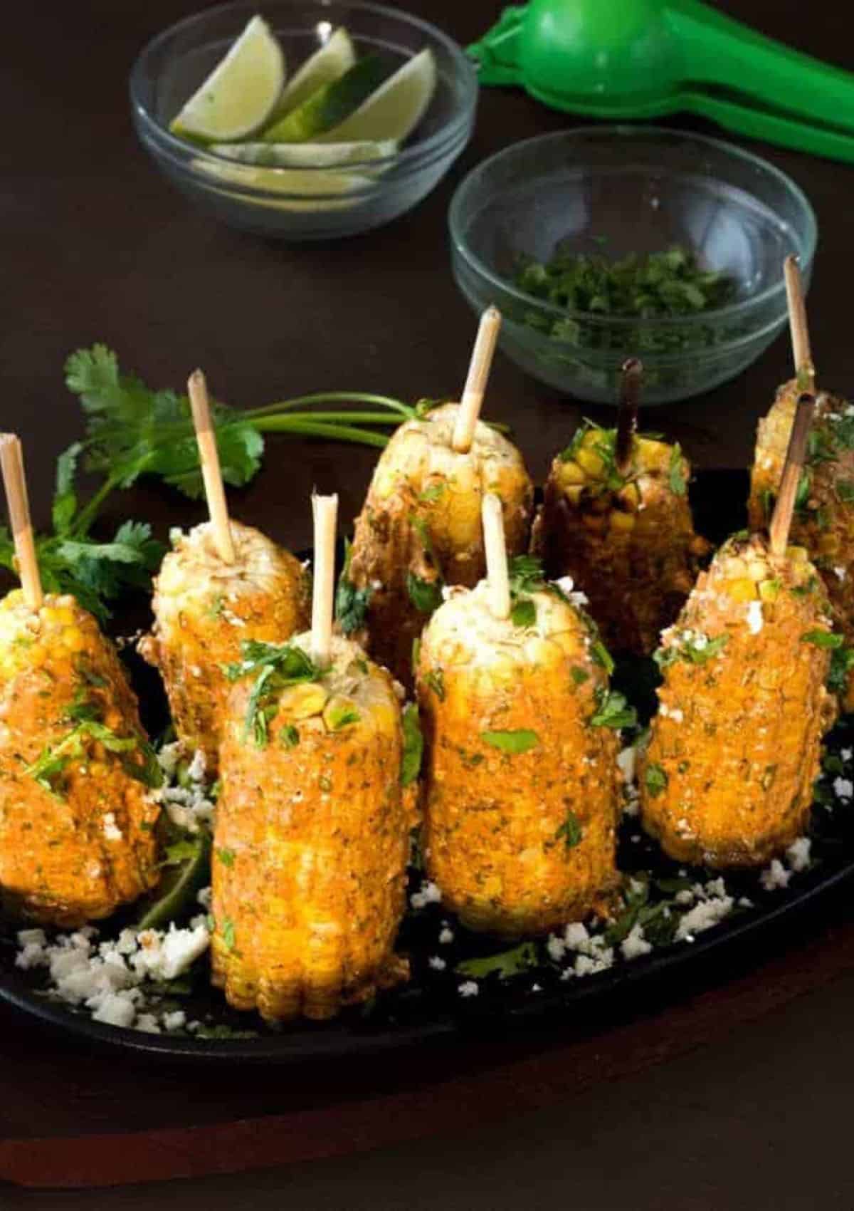 grilled mexican corn on the cob on sticks on a black plate.