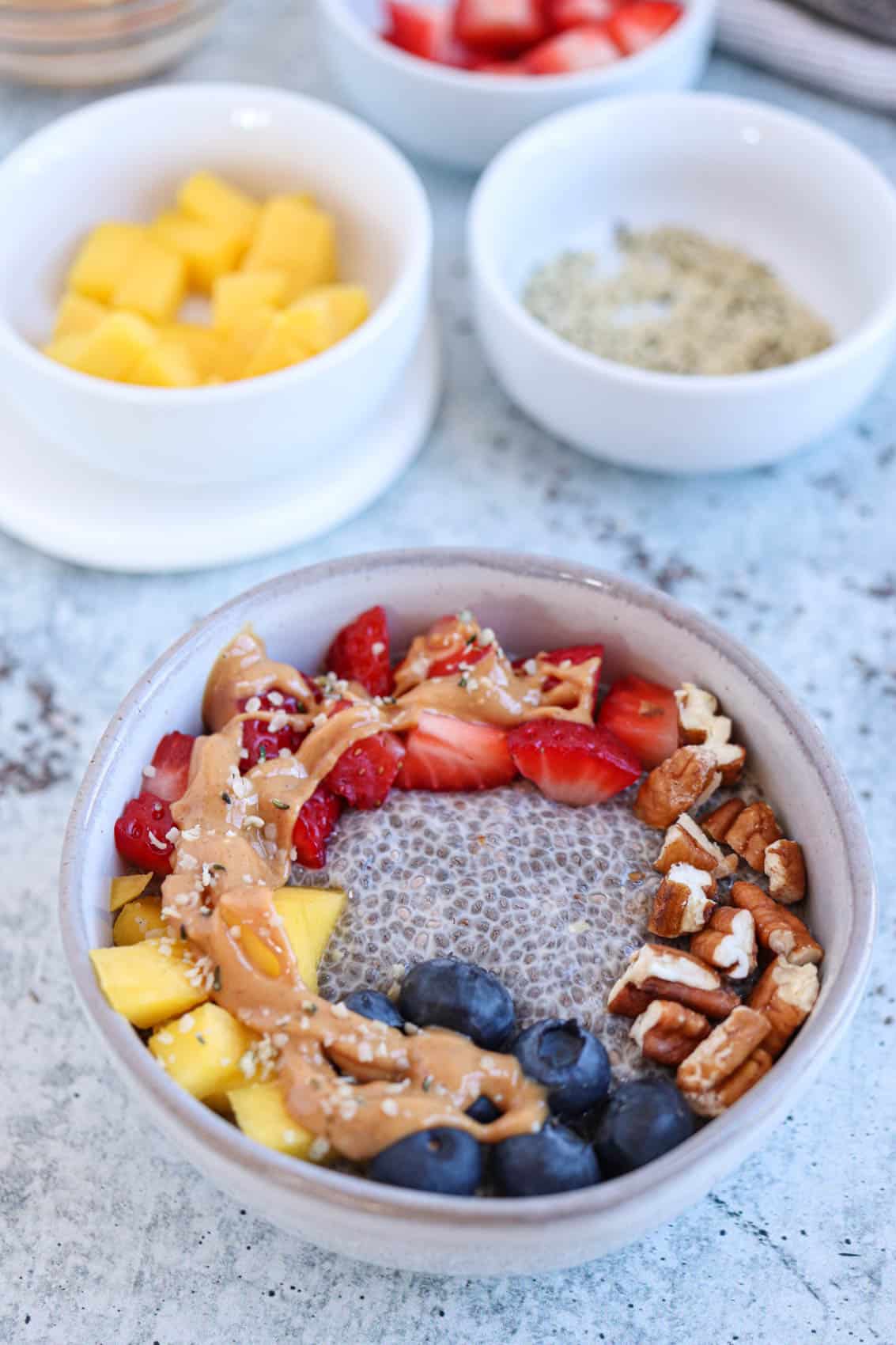 gray bowl with chia pudding, fresh fruit, nut butter and hemp hearts on a light gray surface. 