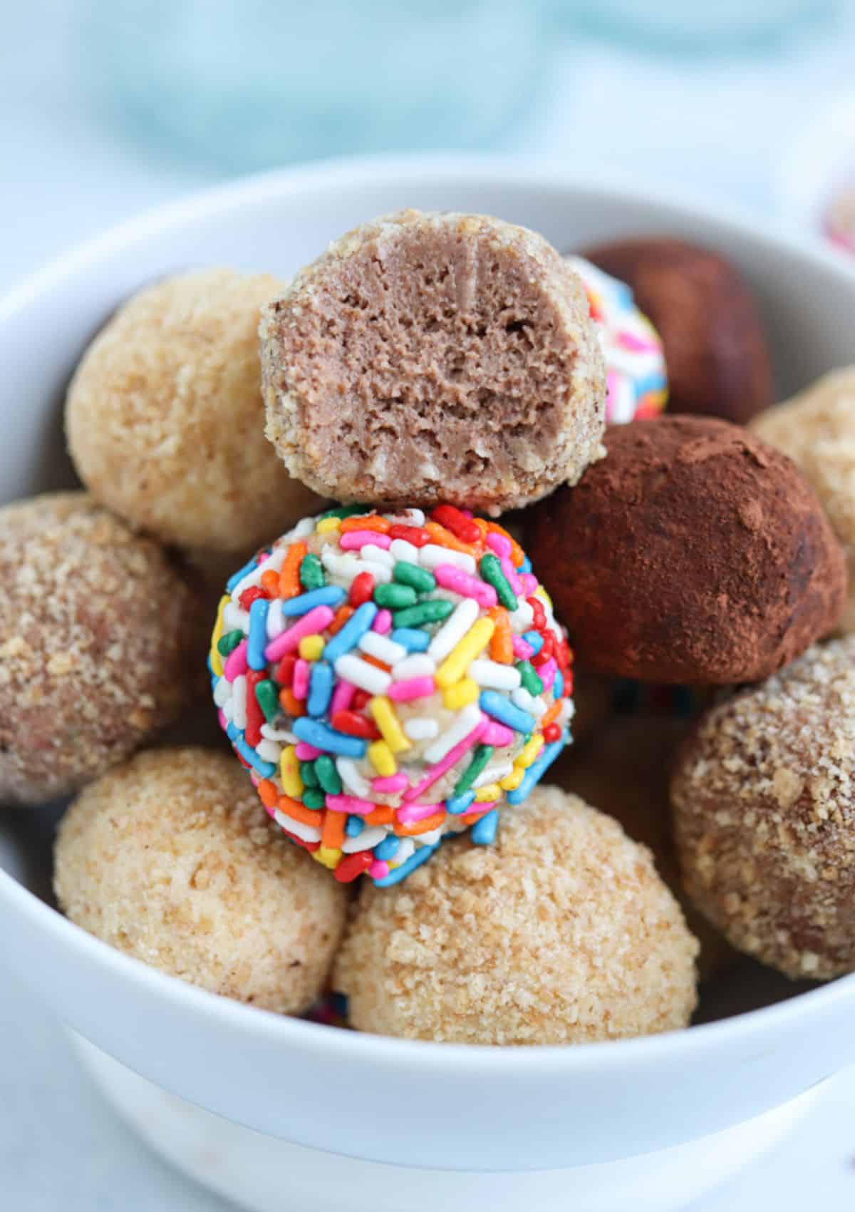 cheesecake balls coated in rainbow sprinkles, cookie crumbs and cocoa powder in a bowl.