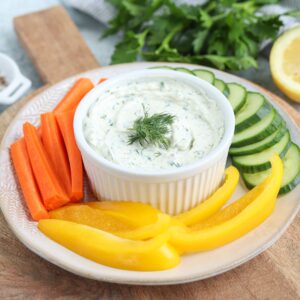 A plate with carrot sticks, cucumber slices, and yellow bell pepper strips surrounds a bowl of cottage cheese ranch dip garnished with dill, with fresh herbs and a lemon in the background.