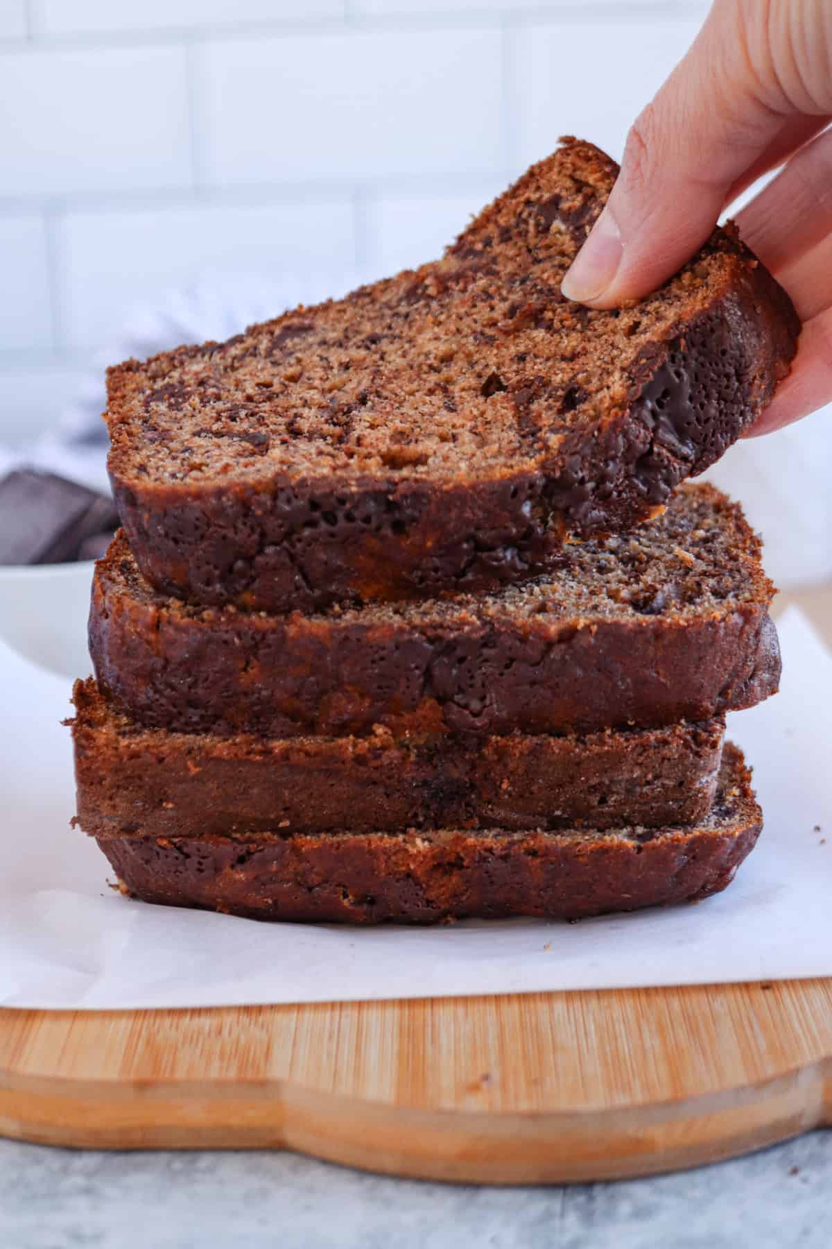 A hand lifts a slice from a stack of banana bread slices, displayed on a wooden board. The breads moist texture and chocolate speckles stand out against the blurred kitchen backdrop.