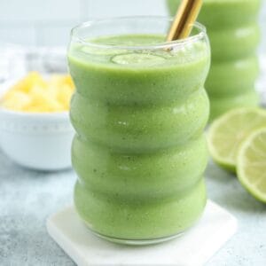 A glass of thick green smoothie with a gold straw, placed on a white coaster. Sliced lime and a bowl of pineapple chunks are in the background, creating a fresh vibe reminiscent of pesto chicken parmesan’s vibrant flavors. The smoothie is in a wavy glass.