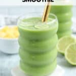 A refreshing pineapple cucumber smoothie in a textured glass with a gold straw, garnished with cucumber slices. In the background, a bowl of pineapple chunks and sliced limes enhances the tropical vibe.