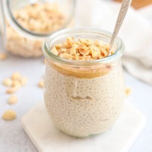 A glass jar filled with chia pudding, topped with chopped nuts and a layer of nut butter, with a spoon inside. Another jar and scattered nuts are in the blurred background.