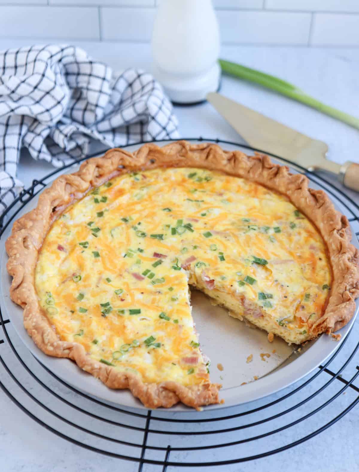 A baked cottage cheese quiche with a golden crust and sprinkled chives sits on a cooling rack, with one slice missing. A kitchen towel, knife, and green onion are in the background.