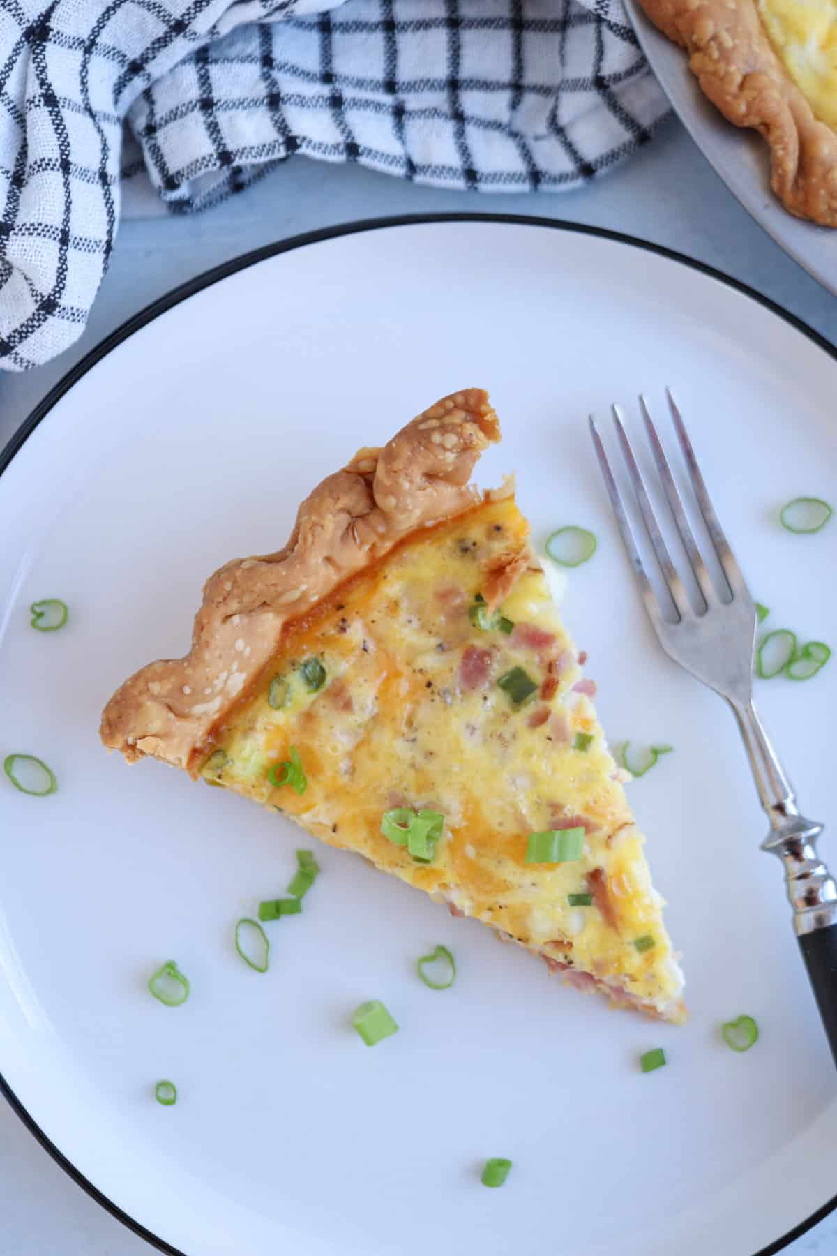 A slice of cottage cheese quiche with a flaky crust sits on a white plate, garnished with chopped green onions. A fork rests beside the quiche, and a black-and-white towel is partially visible in the background.