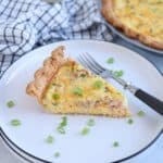 A slice of quiche with diced ham and green onions sits on a white plate with a fork. Chopped green onions are sprinkled around the quiche, evoking the homestyle comfort of a chicken potato broccoli casserole in every bite.