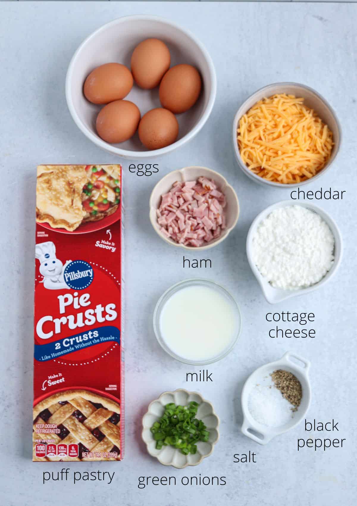 Ingredients for a recipe arranged on a white surface, including eggs, shredded cheddar, diced ham, cottage cheese, milk, black pepper, salt, sliced green onions, and a box of Pillsbury pie crusts—perfect for a cozy chicken potato broccoli casserole.